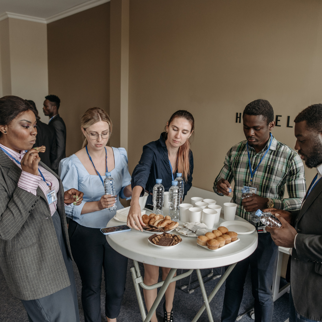 Group of professionals networking casually around a snack table with water bottles and pastries during a business event.